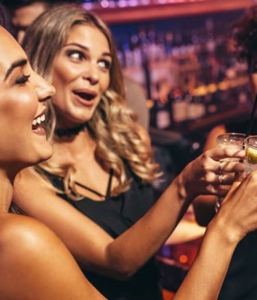Group of happy women laughing and holding cocktails at a crowded bar during a night out.