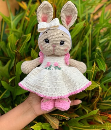 Handmade amigurumi bunny doll wearing a white crochet dress and pink shoes outdoors.