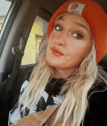 Blonde woman wearing an orange Carhartt beanie and black sweatshirt sitting in a car.