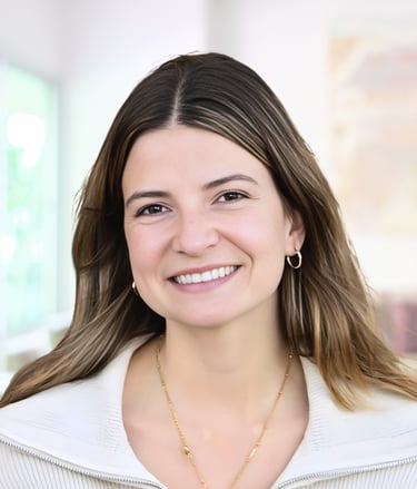 A smiling professional woman with brown hair and gold jewelry wearing a white blazer in an office.
