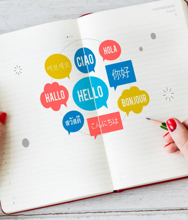 A person writing in an open notebook with colorful speech bubbles showing greetings in multiple languages.
