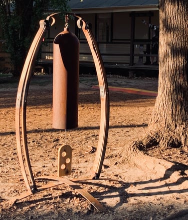 Rustic metal garden bell sculpture hanging on a curved steel frame in a sunset outdoor yard setting.