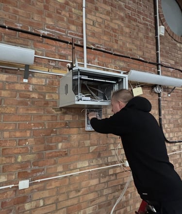 An IT technician installs network cabling and server equipment in a wall-mounted rack on a brick wall.