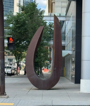 A large oval metal sculpture stands on a modern city sidewalk in Calgary depicting Identity & Worth!