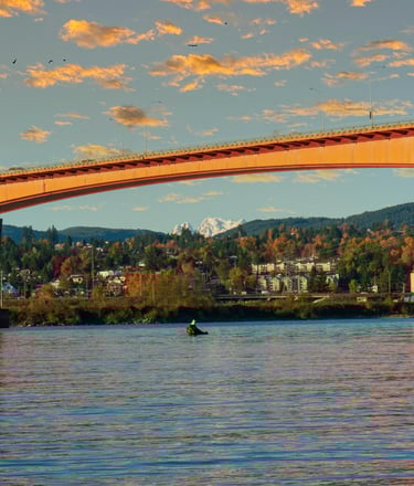 Golden Ears Bridge spanning a river, with houses and autumn-colored trees in the background, and distant snow-capped mountain