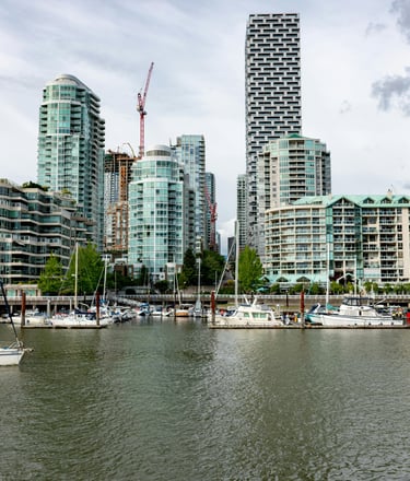 A waterfront scene with high-rise buildings, including some under construction, and sailboats docked along the shore.