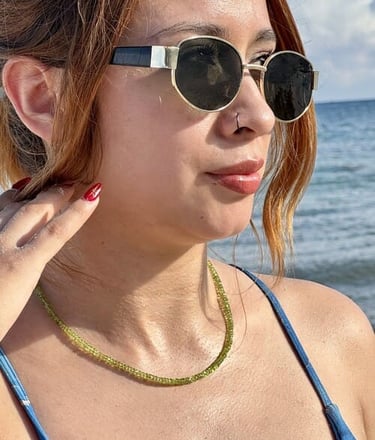 Mujer usando un delicado collar de cuentas de peridoto y gafas de sol redondas en la playa.