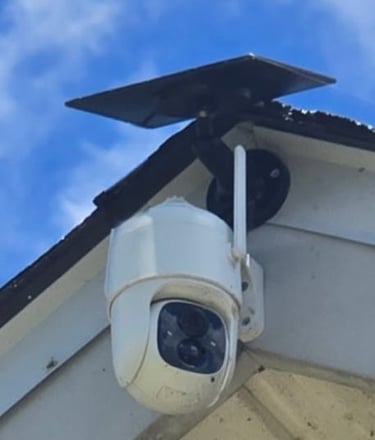 Solar powered wireless outdoor security camera mounted on a house roof against a blue sky.