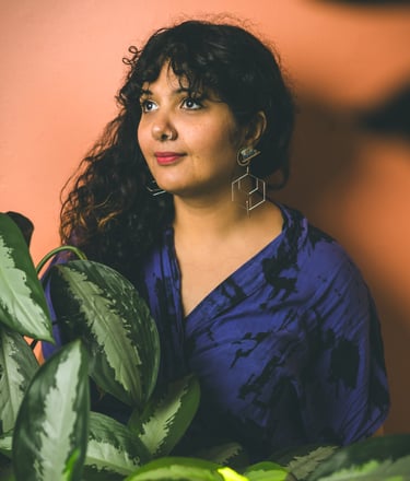 Portrait of Salima a brown skinned woman with long curly dark hair surrounded by plants. 