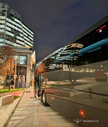 Luxury charter bus parked at night near a modern glass office building with city light reflections.