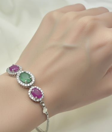 Elegant silver bracelet featuring oval emerald and ruby gemstones with diamond halo settings on a wrist.