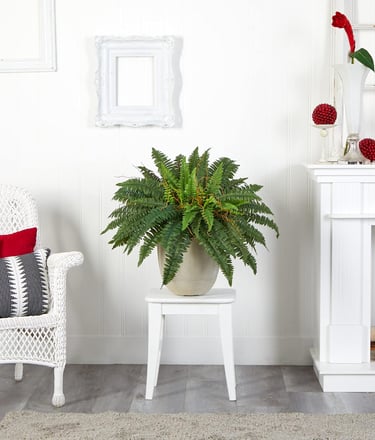A lifelike artificial fern plant in a ceramic pot on a white stool, enhancing a cozy living room with a wicker chair and