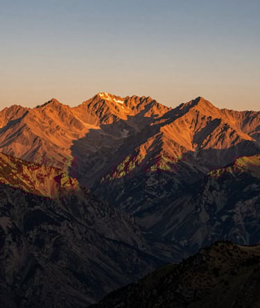 A breathtaking landscape shot of a mountain range at golden hour. The peaks are bathed in warm orange light, with charcoal shadows in the valleys. The composition is authentic and vast.