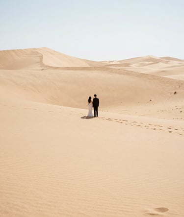 A wide-angle landscape photograph of an elopement in a North American desert. The couple is a small focal point against vast, clean lines of sand dunes. Bright, airy lighting, high-end professional photography style.
