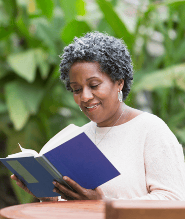 Une femme souriante lisant un livre bleu dans un jardin verdoyant, symbolisant la paix.