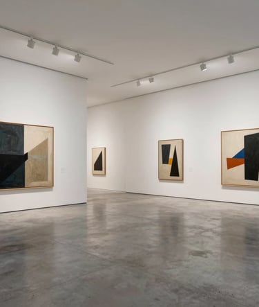 A wide, expansive view of a minimalist visual arts gallery in a European / French city. Large-scale contemporary paintings are displayed on soft off-white walls. The flooring is polished concrete in silver grey. Refined and quiet atmosphere.