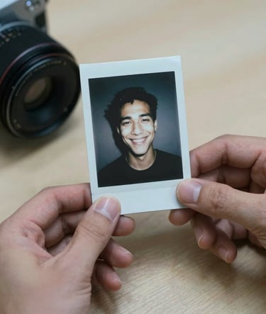 A close-up shot of a pair of hands holding an old polaroid photo of a smiling face. The background is a soft beige wooden surface. The lighting is slightly moody with muted slate blue shadows.