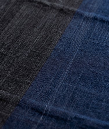 An extreme close-up of textile texture showing the weave of hand-loomed silk. The colors transition from charcoal black to deep indigo, highlighting the organic imperfections of the fiber in a East Asian / South Asian influenced for a global luxury audience design house.