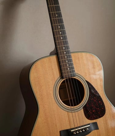 A close-up photograph of a vintage acoustic guitar leaning against a soft taupe wall in a North American / US music room. The lighting is soft and natural, highlighting the wood grain and strings. Moody dark charcoal brown shadows add depth.