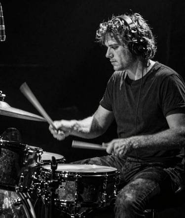 A high-contrast black and white photograph of a drummer in motion during a live set. The lighting is dramatic, capturing the energy of the performance in a Western European / Dutch music venue. Deep black shadows and sharp light grey highlights.