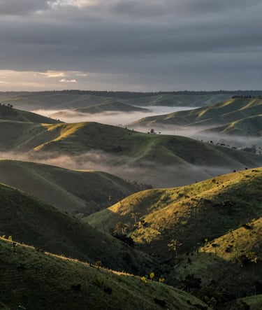 A wide cinematic landscape of the Angolan highlands at dawn, deep slate grey mists rolling over green hills with muted gold sunlight piercing through, high-end documentary photography style.