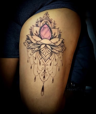 Tatuaje ornamental de flor de loto en el muslo, con una gema rosa y detalles de joya.