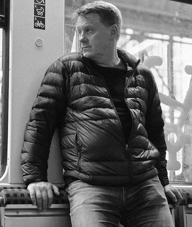 A man in a puffy jacket and jeans leans on a train seat, gazing out the window. Accessibility signs are above. Black and whit