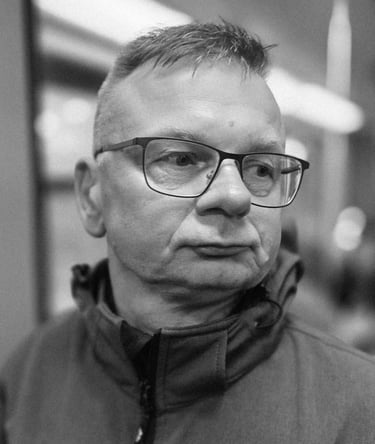 A middle-aged man with short hair and glasses looks sideways indoors, wearing a zip-up jacket. Black and white, blurred backg