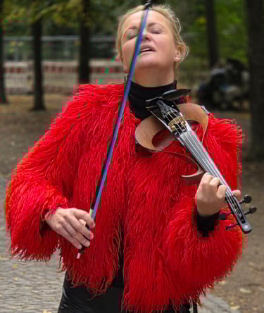 A woman in a bright red shaggy jacket plays her violin outdoors, eyes closed, head back, trees and cobblestones around her.