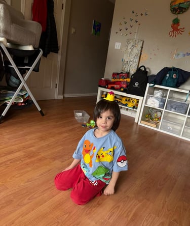 A young boy in a Pokemon shirt sits on a wooden floor in a playroom.
