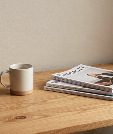 An inspiring home studio workspace featuring a warm light wood desk, a premium ceramic mug, beautiful design magazines stacked neatly, and a soft beige linen background, North American / Modern Western.