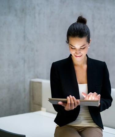 A businesswoman in a black jacket, sitting on a desk, holding a tablet computer.