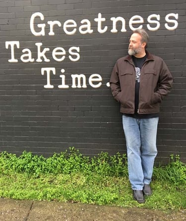 Jason leaning against a wall with a painted message that greatness takes time