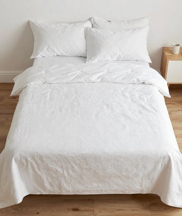 Minimalist white bedding set with textured duvet cover and pillows on a wooden floor.