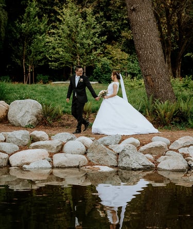 A newlywed couple walks hand-in-hand by a reflective pond during an outdoor garden wedding ceremony.
