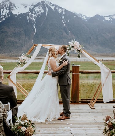 A bride and groom sharing their first kiss under a wooden geometric arch during an outdoor mountain wedding ceremony.