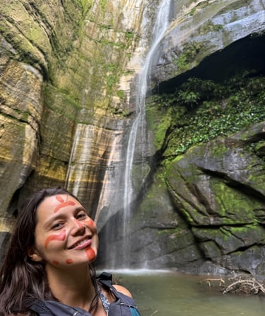 Une femme souriante, le visage peint selon la tradition indigène, se tient devant une haute cascade