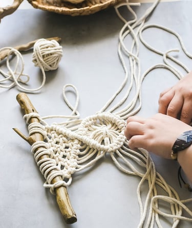 Manos elaborando un tapiz de macramé con cordón de algodón blanco sobre una rama de madera natural.