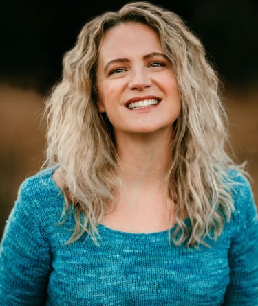 A smiling woman with wavy blonde hair wearing a blue knit sweater in a natural outdoor setting.