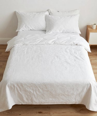 Minimalist white bedding set with textured duvet cover and pillows on a wooden floor.