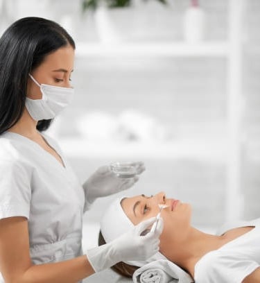 Professional esthetician in a mask applying a chemical peel facial treatment to a client in a spa.