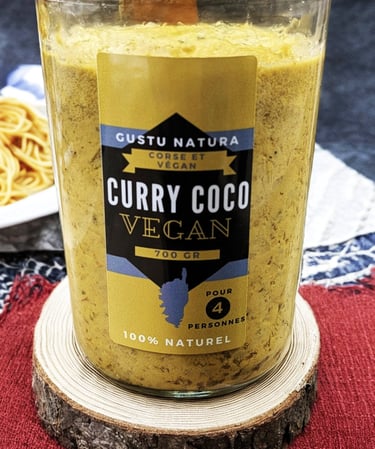 Glass jar of Gustu Natura vegan curry coco sauce on a rustic wooden coaster with pasta background.
