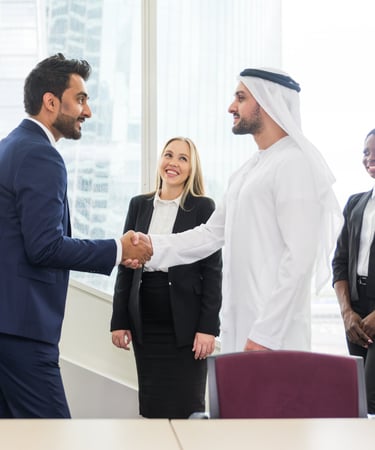 Businessman in a suit shakes hands with another in Emirati attire, while a businesswoman smiles