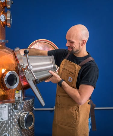 Mastro distillatore che sta caricando il suo alambicco con un secchio di acciaio in mano