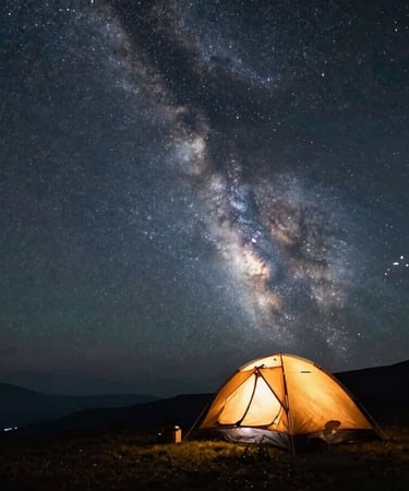Wide starscape with the Milky Way arching over a quiet mountain camp, a single silhouetted tent glowing with warm amber light inside.