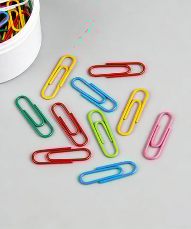 A collection of colorful metal paper clips scattered on a gray surface next to a white container.