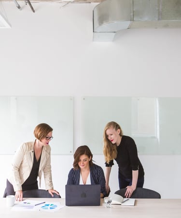 three women standing around a table with laptops