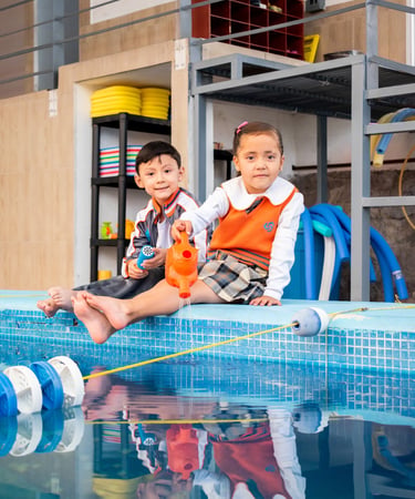 Two school children in uniforms sitting by an indoor swimming pool playing with a watering can.