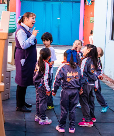 A preschool teacher talks to a group of young students in school uniforms on an outdoor playground.