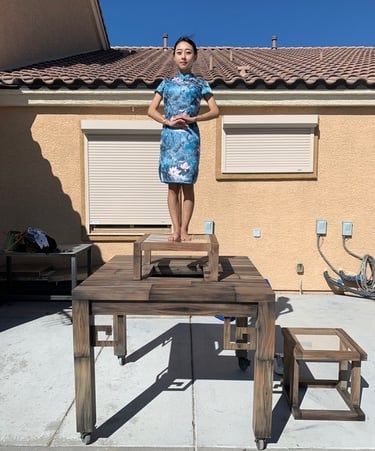 A woman in a blue floral cheongsam dress stands on a tiered wooden table platform outdoors.